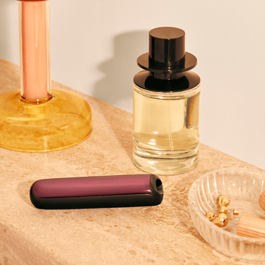 Makyaj masası üzerinde duran cihaza takılı Dark Orchid renkli Ploom AURA ön panel aksesuarı.
