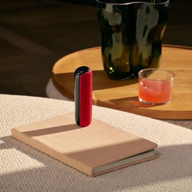Ploom AURA with Lava Red back cover placed on top of a book on a table.
